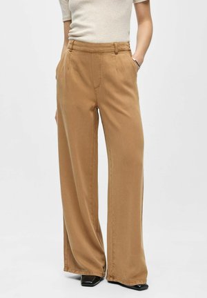 Personne portant un pantalon camel taille haute, jambes larges et un haut beige clair à manches courtes, mains dans les poches.