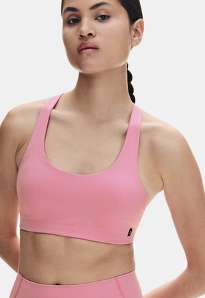 Frau mit geflochtenem Haar, die einen pinkfarbenen Sport-BH und passende hoch taillierte Leggings vor einem schlichten weißen Hintergrund trägt.