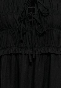 Haut en tissu noir avec sections élastiques froncées et cordons noués en nœud papillon au niveau de l'encolure, montrant la texture et les détails de couture.