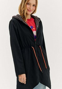 Sieviete valkā melnu jaku ar rāvējslēdzēju un oranžiem auklīšu galiem, ar pārbaudītu kapuci, virs sarkanā krekla ar krāsainu grafiku.