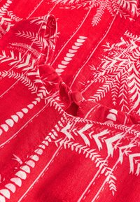 Tissu rouge avec des motifs de fougères blanches, présentant des rayures verticales et une finition texturée. Comprend une section froncée avec un petit détail de bouton.