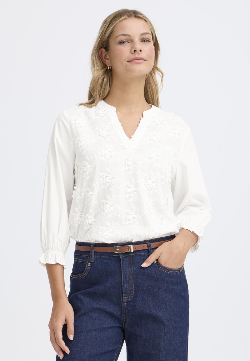 Femme portant un blouse en tissu floral blanc à manches trois-quarts et un jean bleu avec une ceinture marron, se tenant debout avec une main dans sa poche.