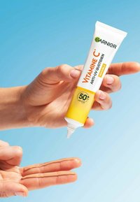 Tube de crème solaire avec un design blanc et jaune, étiqueté "VITAMINE C," affichant un SPF de 50+. Crème solaire liquide sur les doigts, main tenant le tube.