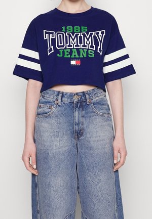 Osoba ubrana w granatowy crop top w białe paski z napisem "1985 TOMMY JEANS", zestawiony z wysokim stanem, jasnoniebieskimi dżinsami.