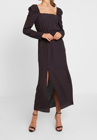 Lång, svart klänning med fyrkantig halsringning, långa ärmar, puffiga axlar, röd prickmönster och en hög slits på sidan. Bärs med svarta sandaler med remmar.