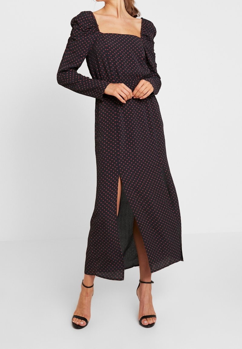 Lång, svart klänning med fyrkantig halsringning, långa ärmar, puffiga axlar, röd prickmönster och en hög slits på sidan. Bärs med svarta sandaler med remmar.