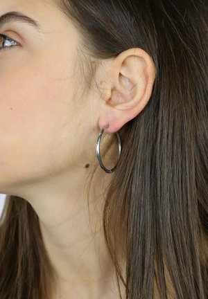 Profilo di una donna con lunghi capelli castani che indossa un orecchino argento a cerchio sull'orecchio visibile, su uno sfondo semplice.