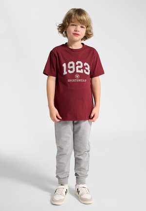 Młody chłopiec o blond włosach, ubrany w bordową koszulkę z napisem "1923 SPORTSWEAR", szare spodnie dresowe i białe trampki, stojący na jednolitym tle.