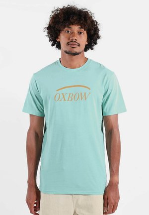 Homme aux cheveux bouclés portant un t-shirt OXBOW turquoise clair et un pantalon beige, debout contre un fond blanc.