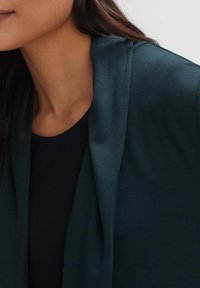 Dark green garment with a smooth texture, draped design, and an open front, paired with a black round-neck top.