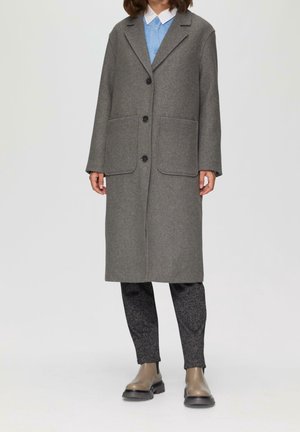 Osoba ubrana w długi szary płaszcz na guziki z dużymi przednimi kieszeniami, ciemne teksturowane spodnie, beżowe buty i niebieską koszulę z kołnierzykiem pod spodem.