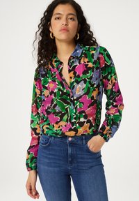 Kleurrijke blouse met een bloemmotief in groen, roze en zwart; heeft een kraag en een knoopsluiting aan de voorkant, gecombineerd met een paar blauwe jeans.
