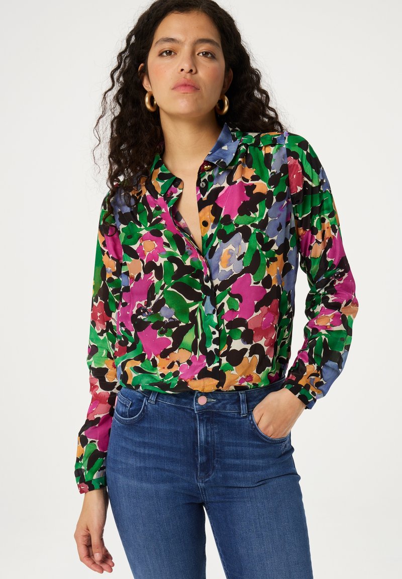 Kleurrijke blouse met een bloemmotief in groen, roze en zwart; heeft een kraag en een knoopsluiting aan de voorkant, gecombineerd met een paar blauwe jeans.