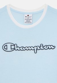 Svijetloplavi pamučni majica s bijelim okruglim izrezom. Na prsima se nalazi istaknut crni "Champion" logo u kurzivnom fontu.