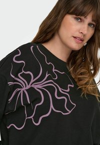 Črna sweatshirt majica z velikim valovitim lila cvetličnim vzorcem na levem ramenu. Tkanina se zdi mehka in ima sproščen kroj.