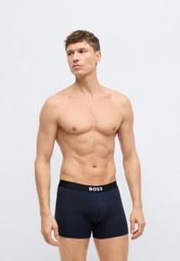 Crni pojas sa logotipom "BOSS", tamnoplava tkanina, uske bokserice, glatke teksture, srednje visoki kroj, nosi ih model atletske građe.