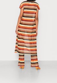 Conjunto multicolor en capas, transparente, con patrón en zigzag en tonos cálidos. Cuenta con mangas cortas, una blusa larga y pantalones acampanados, combinados con tacones naranjas.