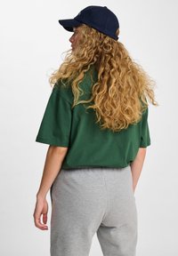 Zielony oversize'owy t-shirt połączony z szarymi dresami, kobieta z kręconymi włosami nosząca granatową czapkę. Prosty, casualowy design z luźnym krojem.