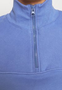 Sweat-shirt bleu clair à col montant, avec une fermeture éclair en plastique et une texture côtelée le long du col et des coutures des épaules.