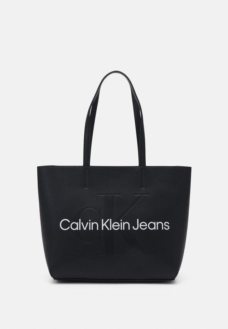 Melnā tote soma, izgatavota no gludas materiāla, ar reljefa "CK" logo un baltu "Calvin Klein Jeans" tekstu priekšpusē. Garas rokturi ērtai nēsāšanai.