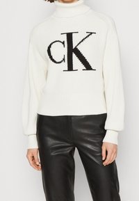 Pull à col roulé blanc côtelé avec un grand logo "CK" noir sur le devant, assorti à un pantalon en cuir noir.