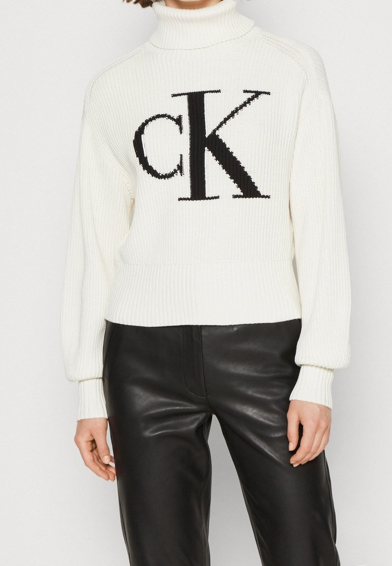 Pull à col roulé blanc côtelé avec un grand logo "CK" noir sur le devant, assorti à un pantalon en cuir noir.