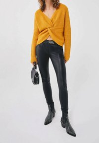 Femme portant un pull jaune moutarde noué à l'avant, un pantalon en cuir noir, des bottines noires cloutées, tenant un sac à main noir.