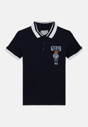 Μπλε πόλο μπλούζα με λευκές ρίγες στο γιακά και τις μανσέτες, με κείμενο "GUESS" και κεντητό αρκουδάκι που φοράει καθημερινά ρούχα στο στήθος.