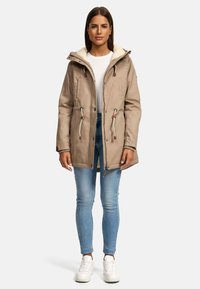 Beige Parka-Jacke mit einem Kragen aus Kunstfell, Zugband in der Taille und zwei Fronttaschen, kombiniert mit hellblauen Jeans und weißen Sneakers.