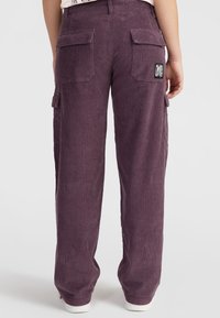 Purple corduroy cargo pants with a relaxed fit, featuring two side pockets and a back patch. Textured material and white sneaker accents.