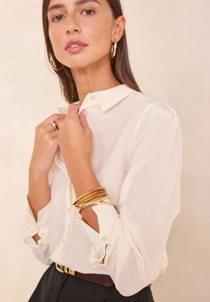 Femme portant un chemisier crème boutonné avec des manches retroussées, des boucles d'oreilles anneaux en or, plusieurs bracelets en or, une bague et un pantalon noir avec une ceinture marron.