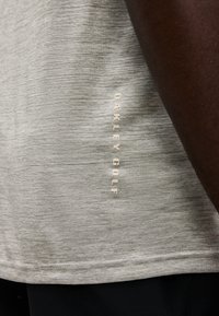 Υφασμα ανοιχτού γκρι με υφή, με κάθετο μπεζ κείμενο "Oakley Golf", μερικώς καλυμμένο από σκουρόχρωμο χέρι στη δεξιά πλευρά.