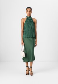 Top a halter plissettato verde abbinato a una gonna in satin coordinata, mentre tiene una piccola borsa rosa. Presenta dei tacchi nere a strappo. Design semplice ed elegante.