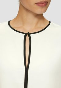Close-up van een vrouw die een wit topje draagt met een zwarte ronde halslijn en een verticale sleutelgat-splijding die is vastgemaakt met een kleine zwarte knop.