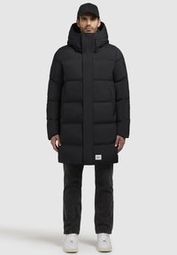 Sort puffet jakke med hætte, der har et quiltet design og en frontlynlås. Båret over grå jeans og hvide sneakers, med et simpelt logo.