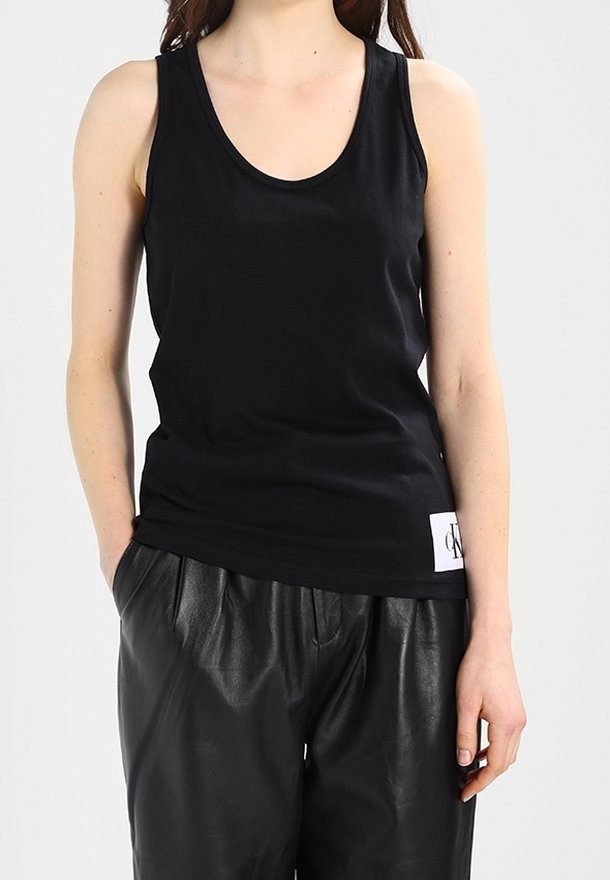 Femme portant un débardeur noir sans manches et un pantalon en cuir noir ample, debout avec une main dans la poche sur un fond uni.