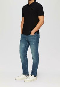 Polo noir avec col et logo doré, associé à un jean bleu clair et des baskets blanches. Accent sur un design décontracté et ajusté.