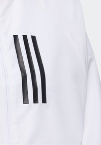 Hvid jakke med en glat tekstur, der har tre lodrette sorte striber på venstre bryst. Lynlås-closure og et sporty design.