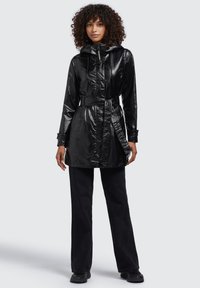 Giacca impermeabile nera e lucida con cappuccio, vita arricciata, chiusura con zip e design con cintura. Presenta maniche lunghe e un orlo leggermente svasato.