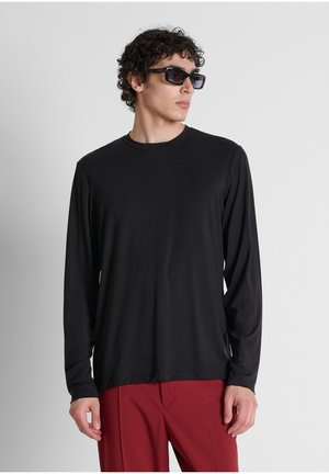 Hombre con camisa negra de manga larga, pantalones rojos y gafas de sol negras, mirando hacia un lado contra un fondo liso gris claro.