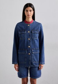 Jeans jakna z zlatimi gumbi, dvema žepoma na prsih in kontrastnim rdečim ovratnikom. Ujemata se z jeans kratkimi hlačami, obema v srednjem pranju.