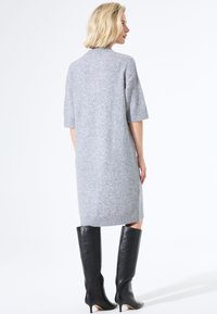 Robe tricotée grise à manches courtes, mi-longue, avec une fente sur le côté. Associée à des bottes en cuir noir à hauteur de genoux avec des talons aiguilles.