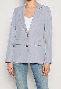 Jasnoniebieska marynarka o dopasowanym kroju, z dwoma przednimi kieszeniami i mosiężnymi guzikami. Wykonana z gładkiego materiału o miękkiej teksturze.