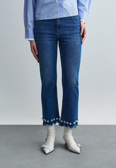Person wearing cropped blue jeans with pearl and flower embellishments at frayed hems, white socks, white pointed heels, and a blue striped shirt.