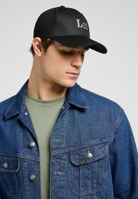 Black cap with embroidered letters "LGC," worn by a model in a denim jacket over a green shirt. The jacket has metal buttons and pockets.