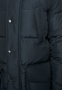 Detailní záběr tmavé prošívané bundy s patentními knoflíky, prošíváním a několika otevřenými i klopovými kapsami na přední straně.