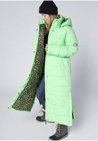 Langer grüner Daunenmantel mit schwarzem Leopardenmuster-Futter, Reißverschluss, verstellbarer Kapuze und seitlichen Schlitzen. Getragen über Jeans und Stiefeln.