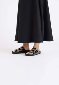 Zwarte sandalen met meerdere banden, voorzien van gouden hardware-accenten, open teenontwerp en rubberen zolen, gecombineerd met een lange zwarte outfit.