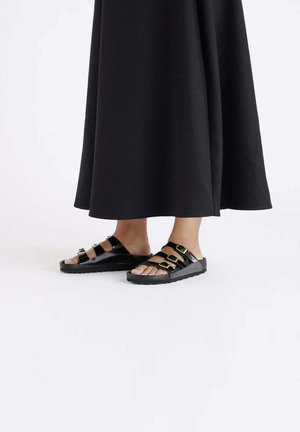 Zwarte sandalen met meerdere banden, voorzien van gouden hardware-accenten, open teenontwerp en rubberen zolen, gecombineerd met een lange zwarte outfit.