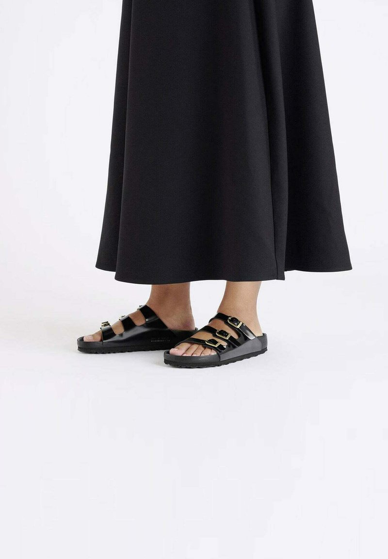 Zwarte sandalen met meerdere banden, voorzien van gouden hardware-accenten, open teenontwerp en rubberen zolen, gecombineerd met een lange zwarte outfit.